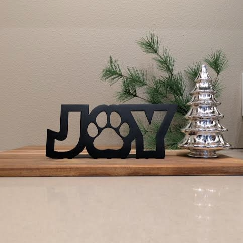 Image of a black metal sign with the word "JOY" and a paw for the "o" from Atomic Rat Rods & Metal Werks. Christmas decorations in the background.