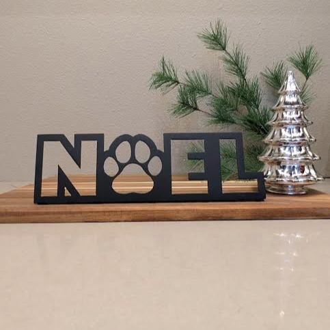 Image of a black metal sign with the word "NOEL" and a paw for the "o" from Atomic Rat Rods & Metal Werks. Christmas decorations in the background.