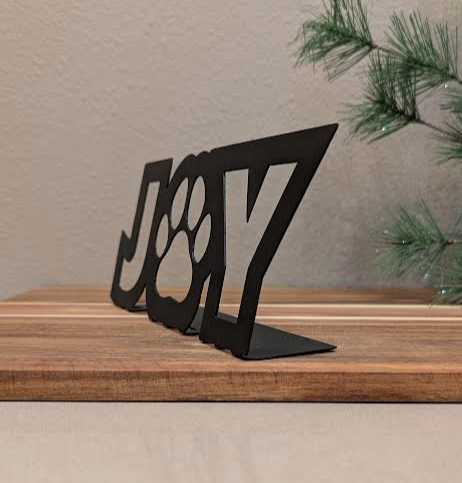 Front corner Image of a black metal sign with the word "JOY" and a paw for the "o" from Atomic Rat Rods & Metal Werks. Christmas decorations in the background.