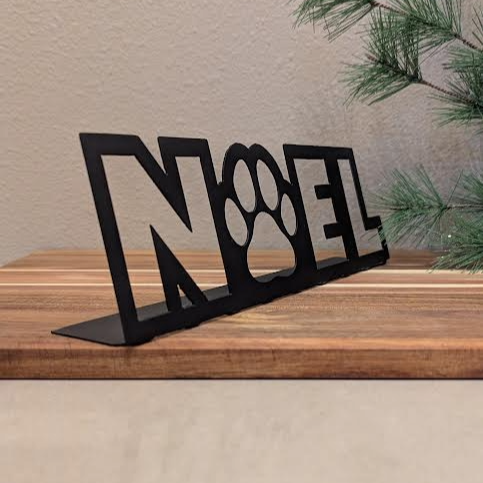 Front quarter view Image of a black metal sign with the word "NOEL" and a paw for the "o" from Atomic Rat Rods & Metal Werks. Christmas decorations in the background.