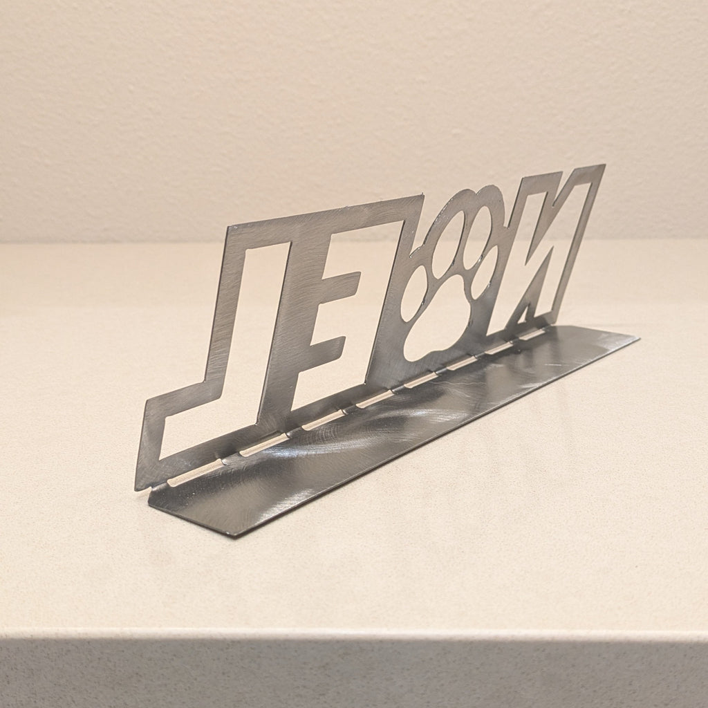 Rear quarter view image of a silver-colored metal sign with the word "NOEL" and a paw in place of the letter "o" from Atomic Rat Rods & Metal Werks.