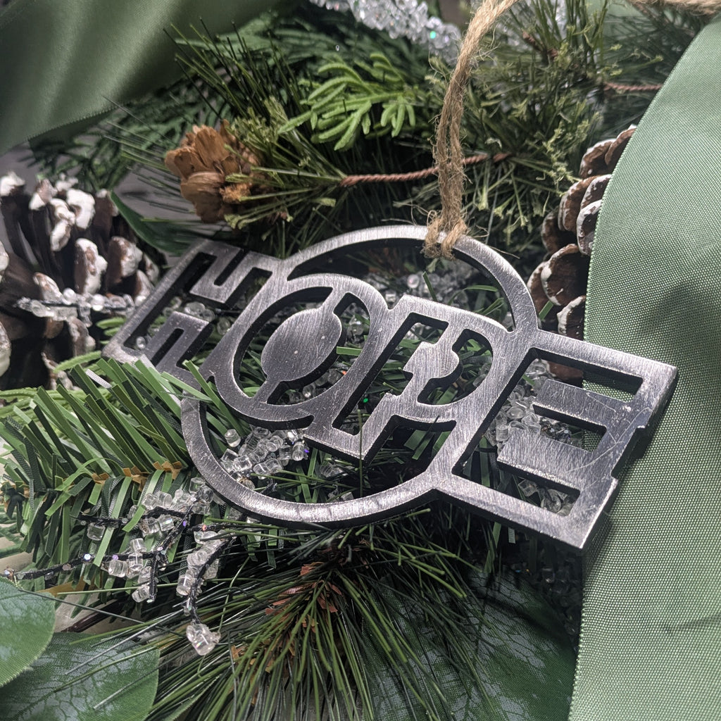 Angle image of a simple, minimalist silver-colored "HOPE" ornament on a Christmas tree from Atomic Rat Rods & Metal Werks.