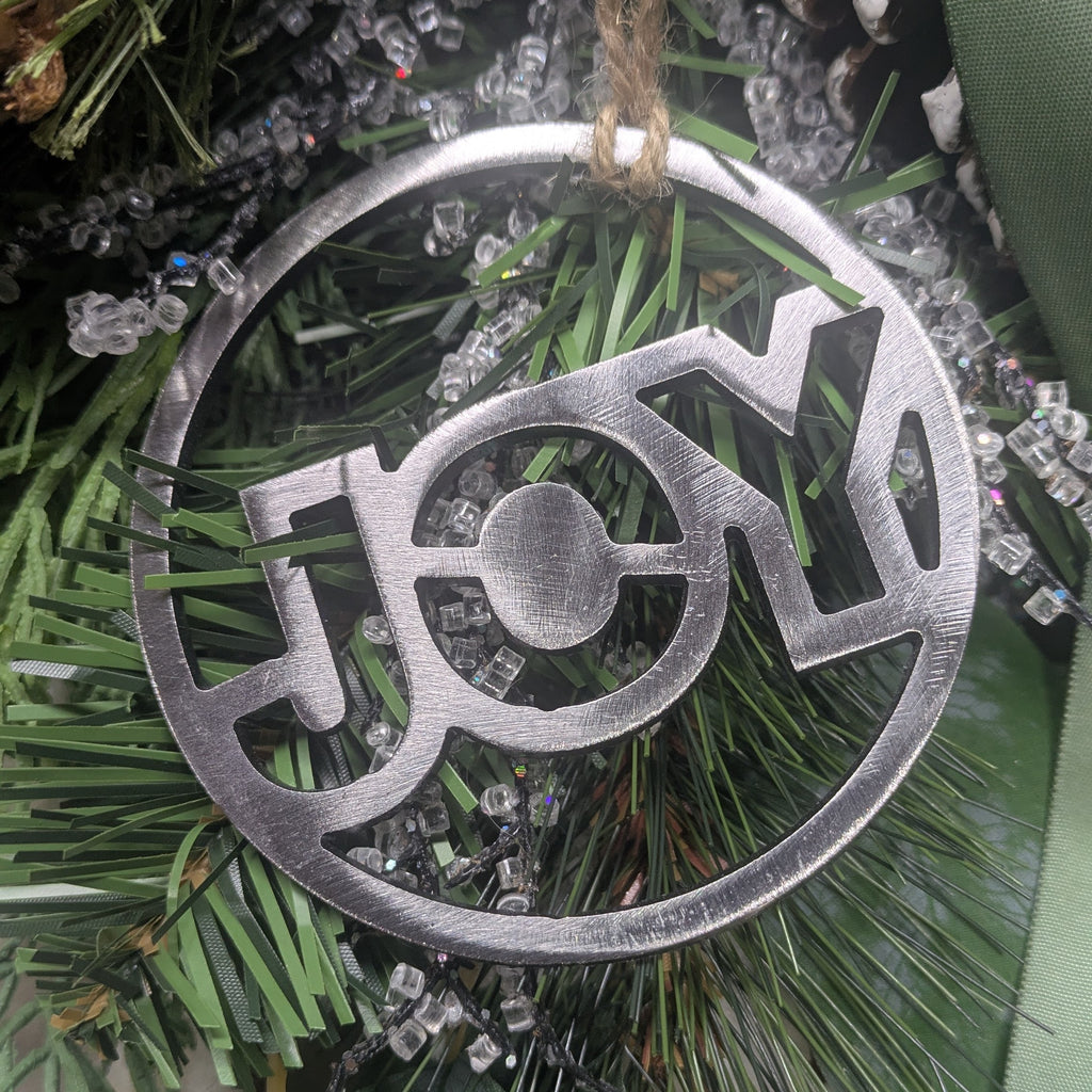 Image of a simple, minimalist silver-colored "JOY" ornament on a Christmas tree from Atomic Rat Rods & Metal Werks.