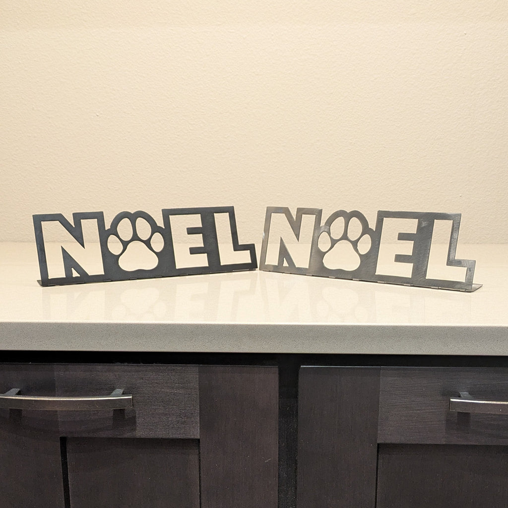 Side by side view of a black metal sign and silver metal sign with the word "NOEL" and a paw in place of the letter "o" from Atomic Rat Rods & Metal Werks.